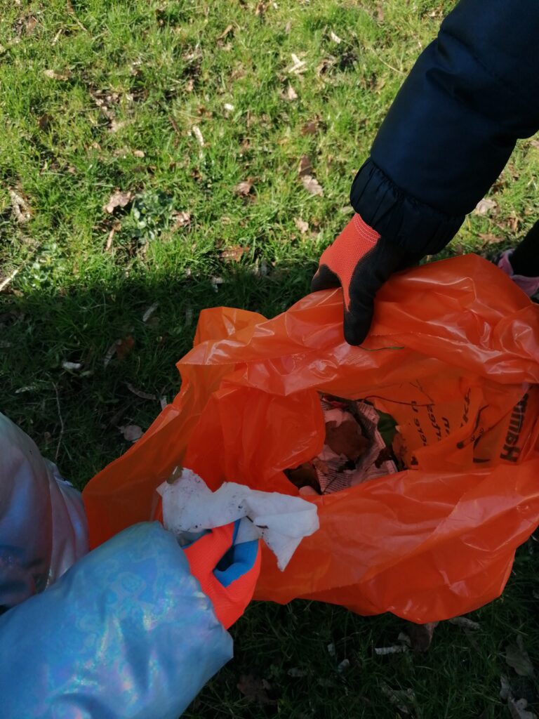 Zwei Kinderhände halten einen orangenen Müllsack. Eine Hand wirft ein benutztes Taschentuch in den Sack.
