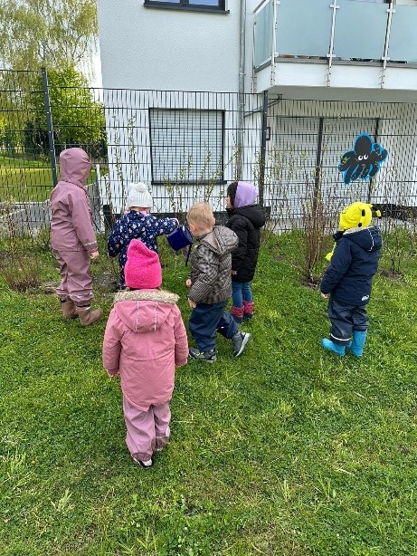 6 Kinder stehen um ein paar kleine Bäumchen, die vor einem Zaun gepflanzt wurden. Ein Kind hat eine Gießkanne in der Hand und gießt ein Bäumchen